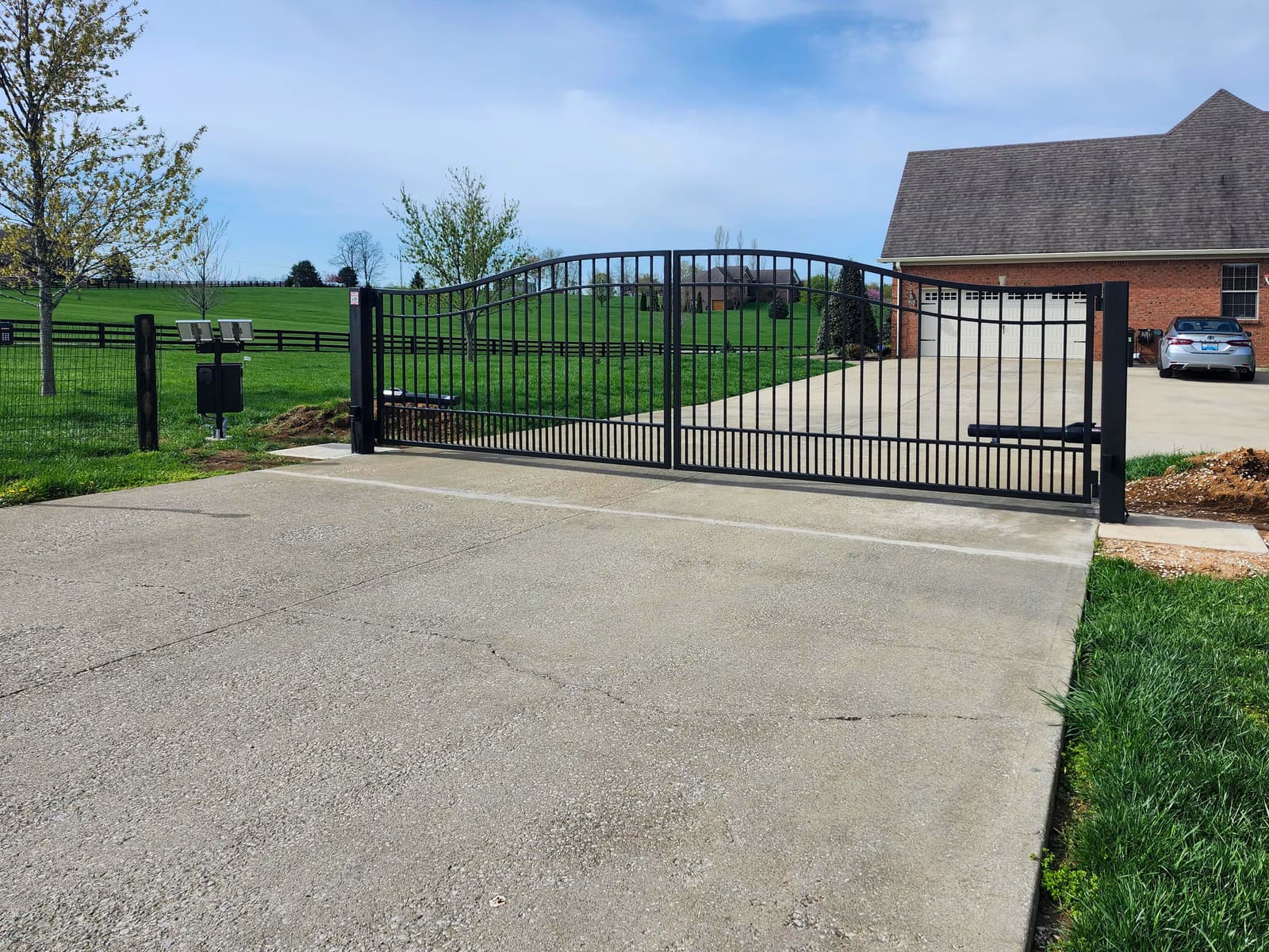 Black arched driveway gate with a solar-powered opener beside a brick home.